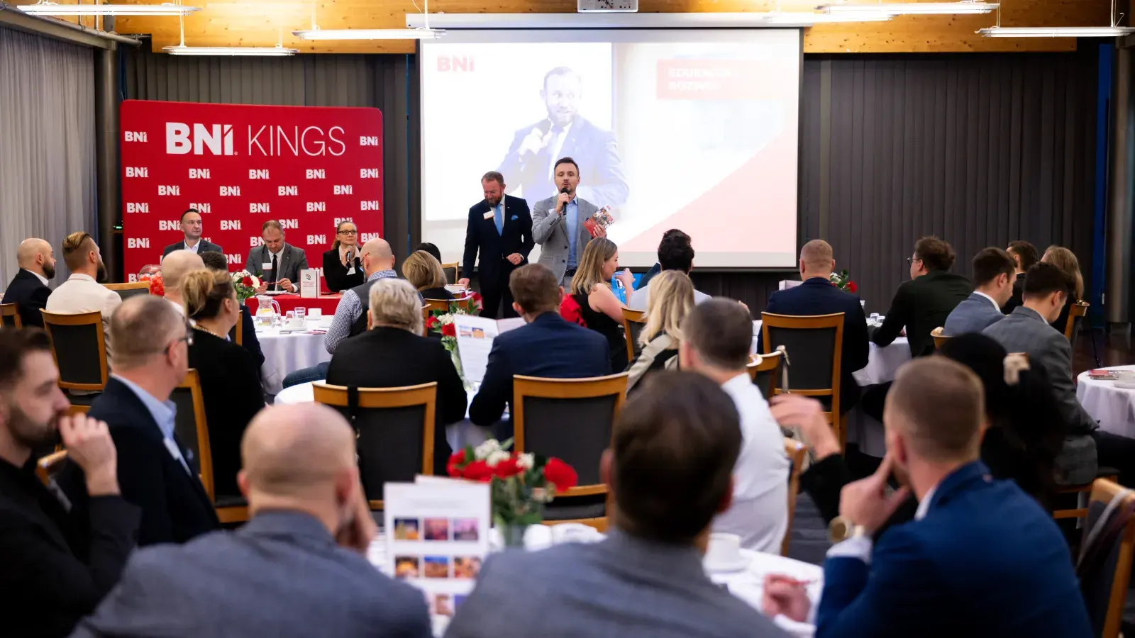 Prowadzący przemawia do uczestników przy stołach, w tle baner Grupy BNI Kings