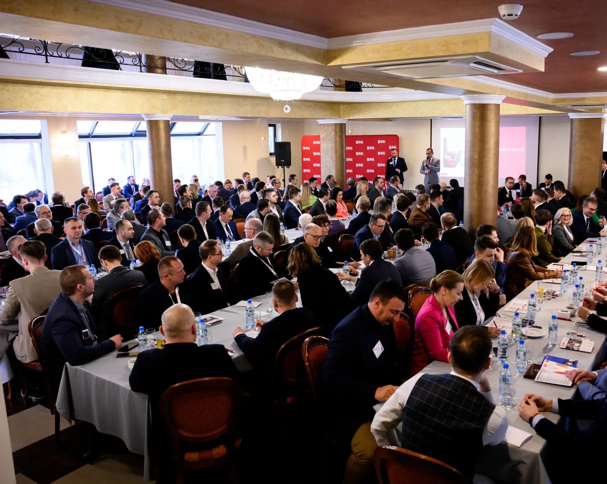 Duża sala z uczestnikami przy długich stołach i roll-upami BNI