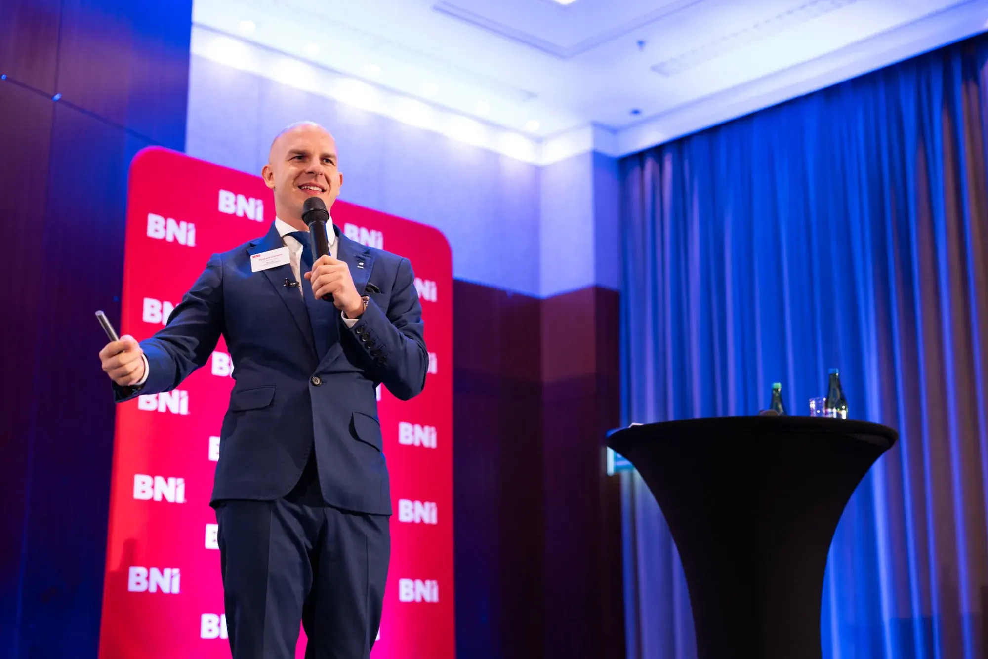 Wystąpienie na scenie BNI z mikrofonem, w tle ścianka z logotypami