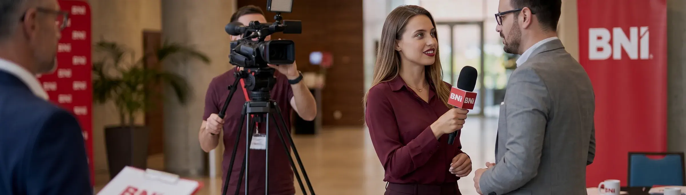 A reporter with a microphone talks to a BNI Member in the lobby of an event; a camera operator stands beside them.