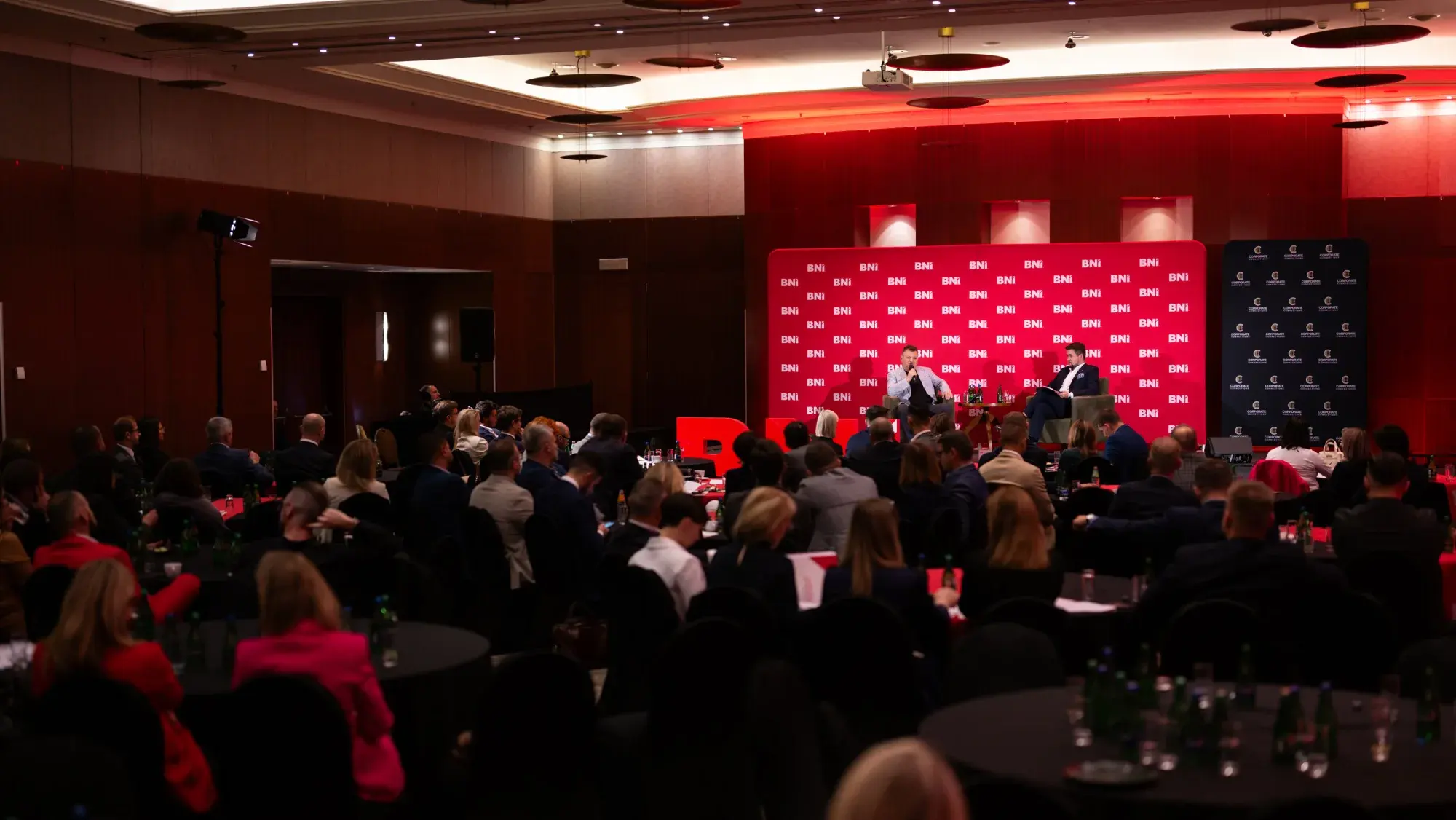 Sala konferencyjna BNI w półmroku, publiczność przed sceną z czerwoną ścianą