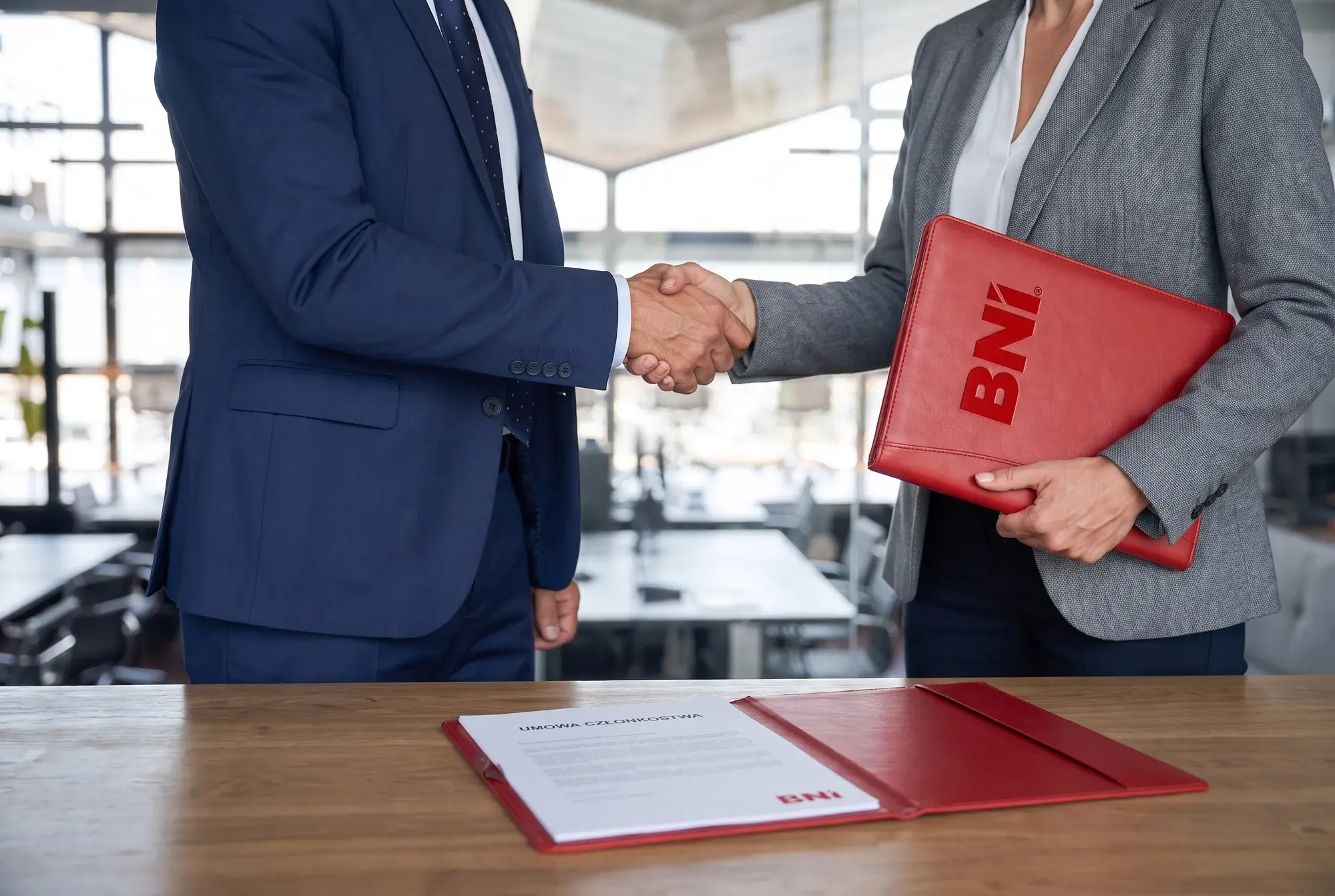 A handshake: a businesswoman holding a red BNI folder greets a business owner