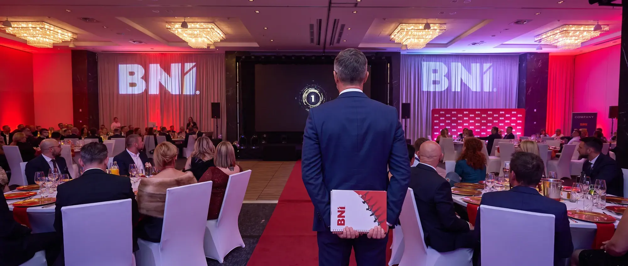 Uczestnik z teczką BNI stoi przed sceną konwencji, obok stoły gości i ekrany z logo BNI
