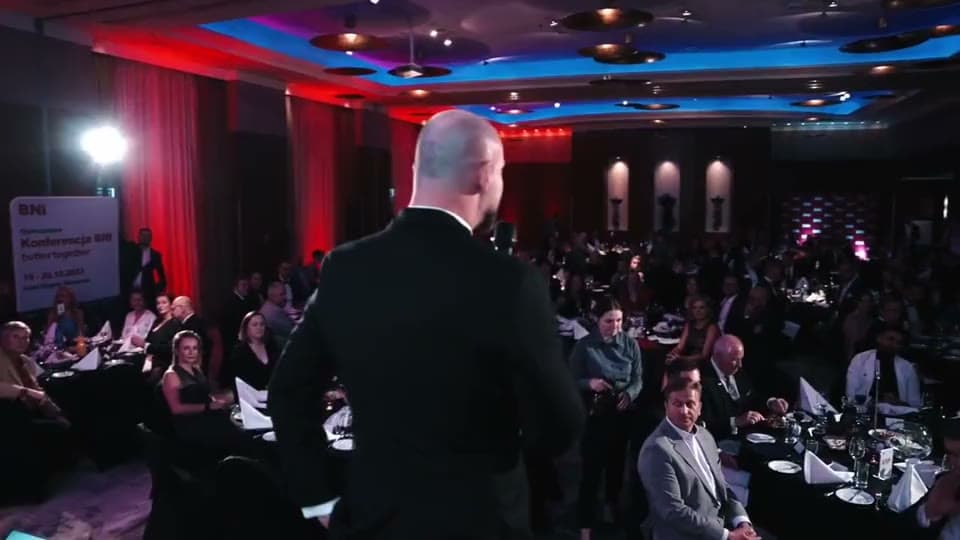 A BNI Polska conference: a full hall at the tables, a speaker on stage by the red BNI wall